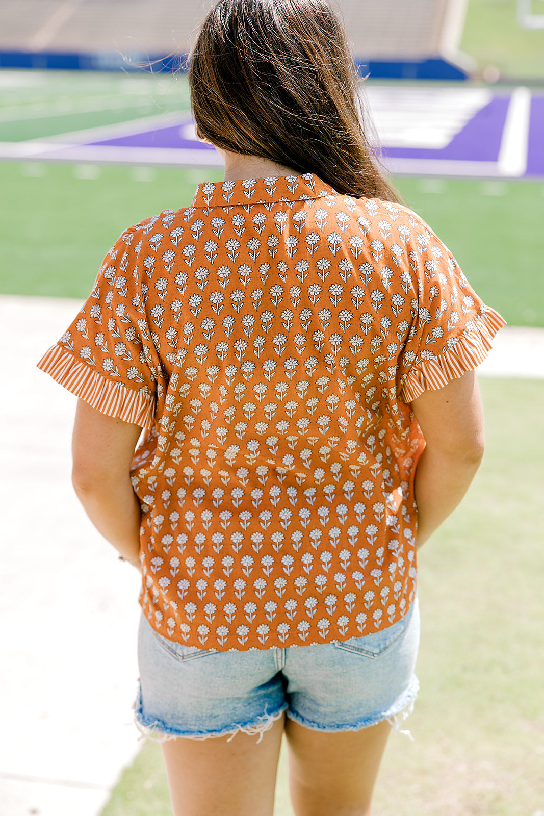 Woman with dark hair facing backwards wearing a short-sleeve, burnt orange top with flower graphics and light-wash denim shorts. 