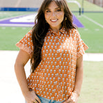 Woman with dark hair facing forwards wearing a short-sleeve, burnt orange top with flower graphics and light-wash denim shorts. 