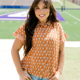 Woman with dark hair facing forwards wearing a short-sleeve, burnt orange top with flower graphics and light-wash denim shorts. 