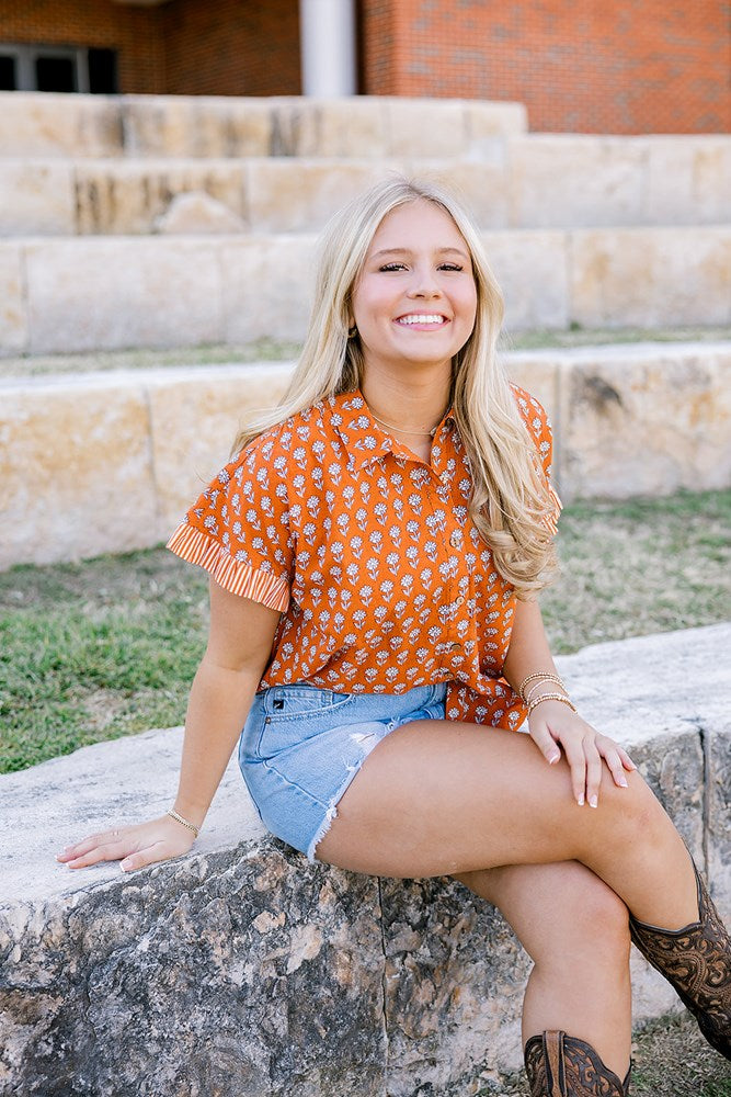 Woman with blonde hair sitting wearing a short-sleeve, burnt orange top with flower graphics, light-wash denim shorts, and boots. 