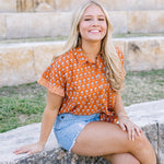Woman with blonde hair sitting wearing a short-sleeve, burnt orange top with flower graphics, light-wash denim shorts, and boots. 
