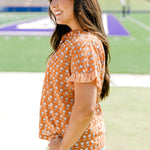 Woman with dark hair facing sideways wearing a short-sleeve, burnt orange top with flower graphics and light-wash denim shorts.