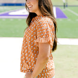 Woman with dark hair facing sideways wearing a short-sleeve, burnt orange top with flower graphics and light-wash denim shorts.