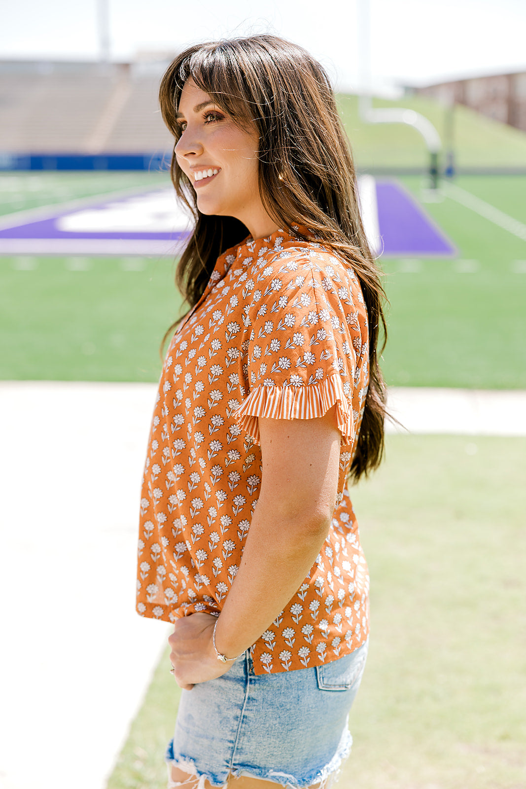 Woman with dark hair facing sideways wearing a short-sleeve, burnt orange top with flower graphics and light-wash denim shorts.