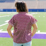 Woman with dark hair standing in a back profile position wearing the Stevie Top in a lifestyle scene. The focal point of this image is the striped ruffle sleeves, and flower graphics on the back of top. The model paired this top with light-wash denim shorts. 