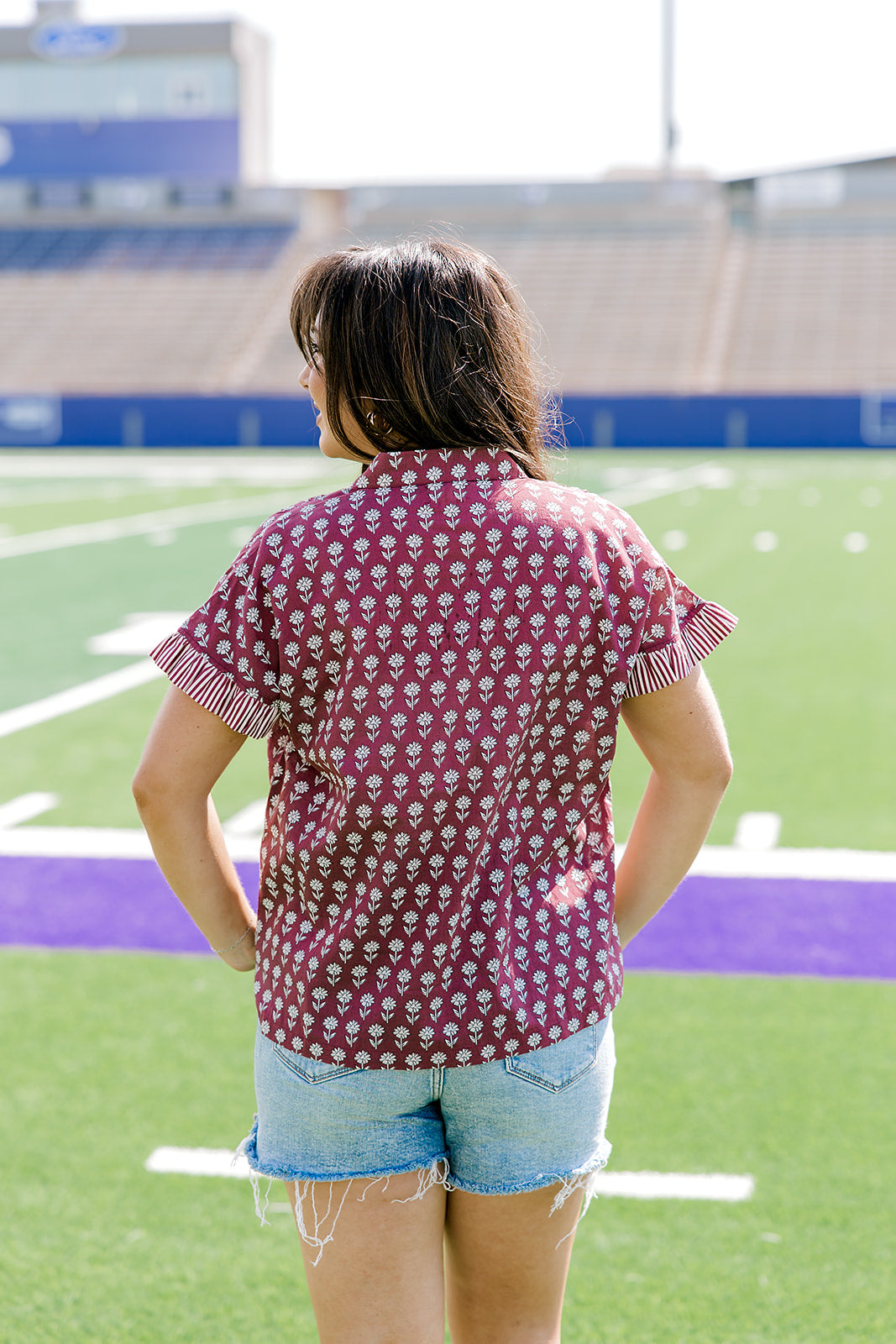 Woman with dark hair standing in a back profile position wearing the Stevie Top in a lifestyle scene. The focal point of this image is the striped ruffle sleeves, and flower graphics on the back of top. The model paired this top with light-wash denim shorts. 