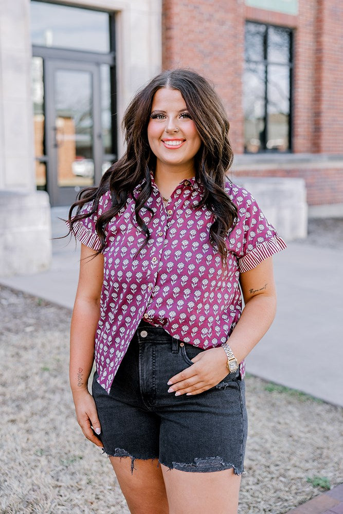 Woman with dark hair standing in a front profile position wearing the Stevie Top on campus. The focal point of this image is the flower graphics on top as well as top in different lighting. The model paired this top with black denim shorts.