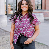 Woman with dark hair standing in a front profile position wearing the Stevie Top on campus. The focal point of this image is the flower graphics on top as well as top in different lighting. The model paired this top with black denim shorts.