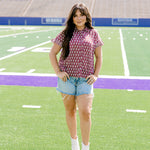 Woman with dark hair standing in a front profile position wearing the Stevie Top in a lifestyle scene. The focal point of this image is the flower graphics on the front of top as well as the wooden buttons down the front. The model paired this top with light-wash denim shorts.