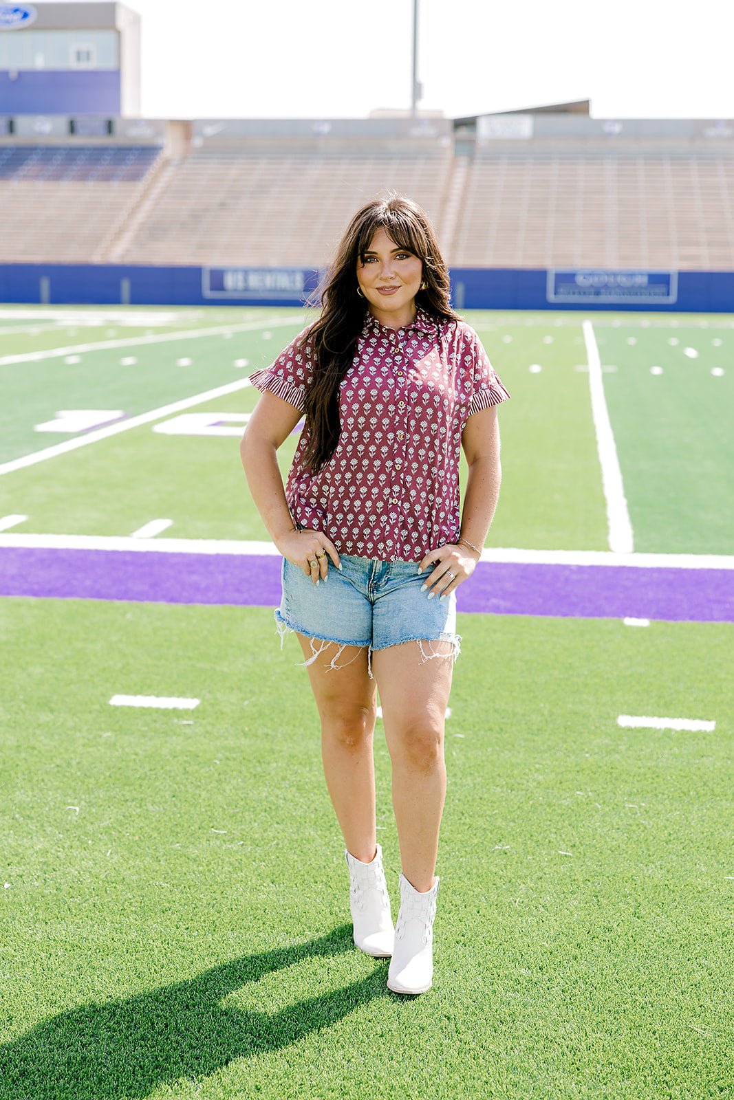 Woman with dark hair standing in a front profile position wearing the Stevie Top in a lifestyle scene. The focal point of this image is the flower graphics on the front of top as well as the wooden buttons down the front. The model paired this top with light-wash denim shorts.