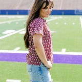 Woman with dark hair standing in a side profile position wearing the Stevie Top in a lifestyle scene. The focal point of this image is the striped ruffle sleeves as well as length of shirt and flower graphics. The model paired this tip with light-wash denim shorts. 