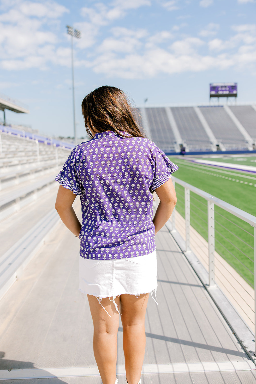 Woman with dark hair standing in back profile wearing the Stevie Top at a football field. The focal point for this image is the graphics on the back of the top as well as the striped ruffle on sleeves. The Model paired this top with a white denim skirt.
