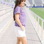 Woman with dark hair standing in side profile position wearing the Stevie Top at a football field. The focal point for this image is the graphics on the side of the top as well as a better view of striping ruffles on sleeve. The model paired this top with a white denim skirt.