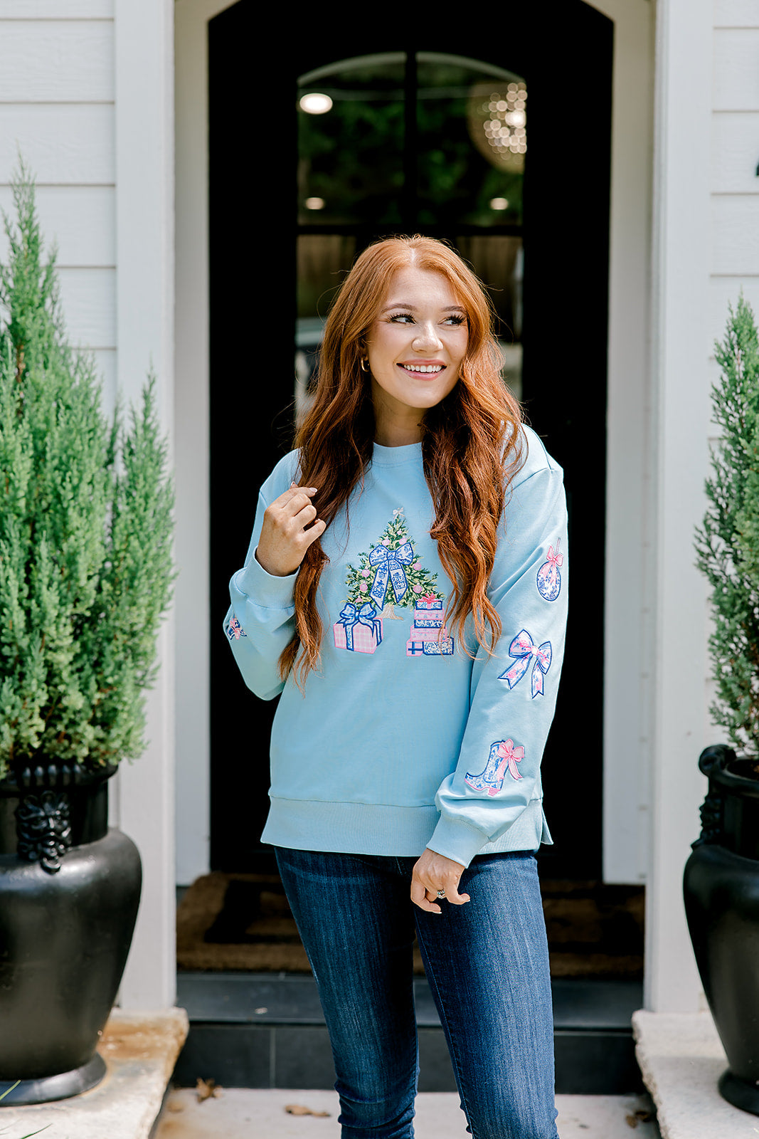 Woman with red hair wearing a baby blue sweatshirt with Christmas motif embroidery. Model paired this top with dark-wash denim jeans. 
