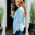 Woman with red hair wearing a baby blue sweatshirt with Christmas motif embroidery. Model paired this top with dark-wash denim jeans. 
