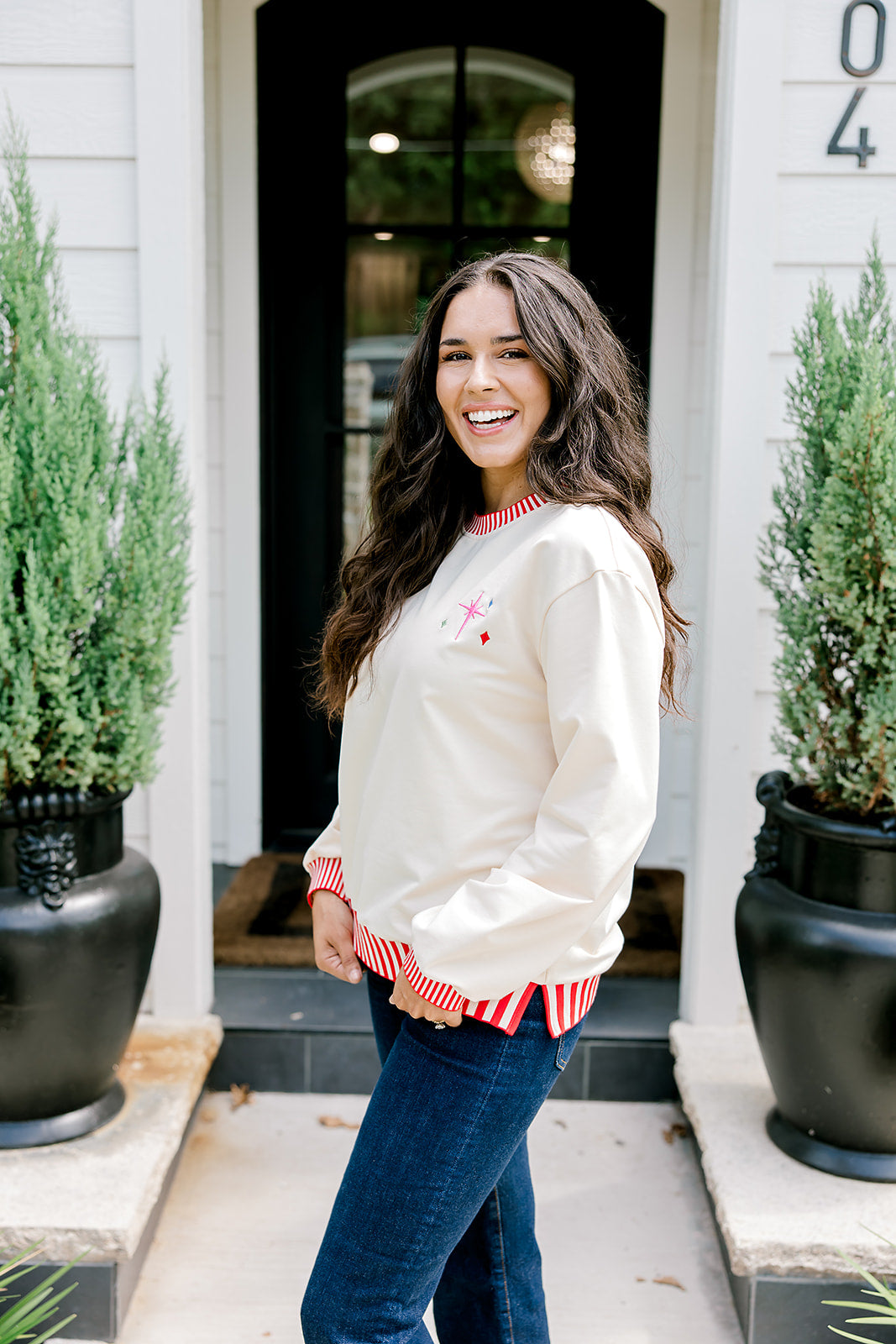 Woman with dark hair wearing a sweatshirt with red & white striping along cuff and collar. On the back of top there is a multi color bible verse. Model paired this top with dark-wash denim jeans. 