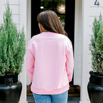 Woman with dark hair wearing a baby pink sweatshirt with embroidered Christmas motifs on front and sleeves of top. Model paired this top with light-wash denim jeans. 