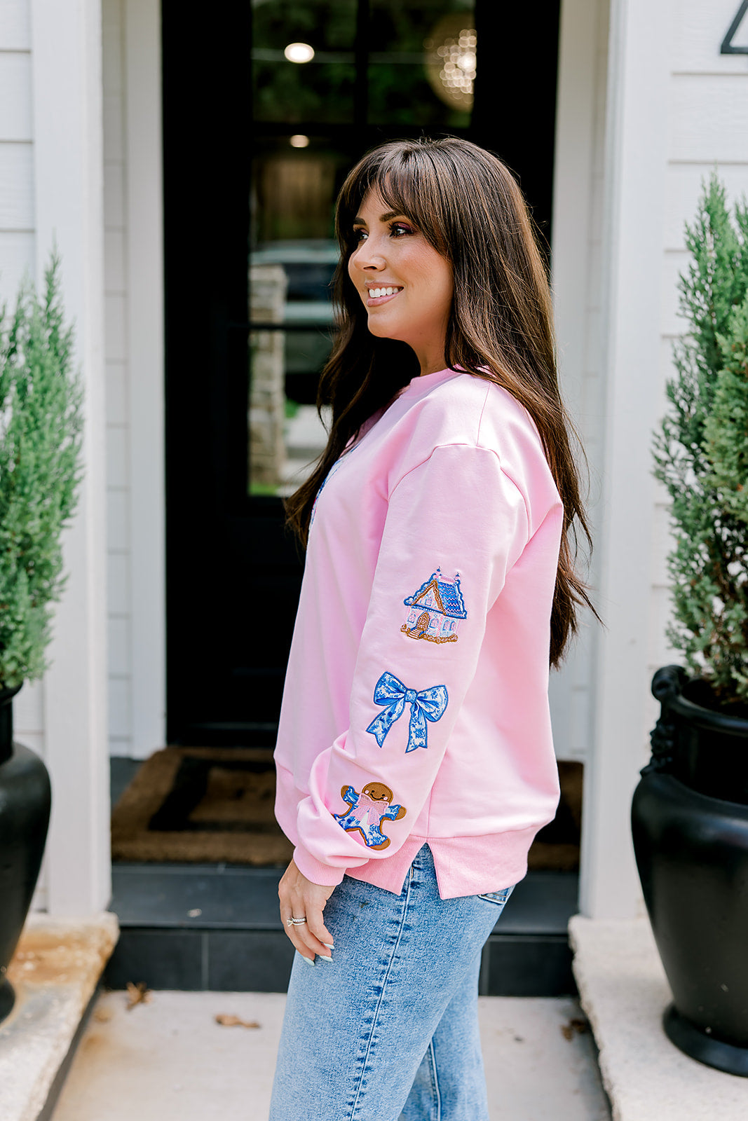 Woman with dark hair wearing a baby pink sweatshirt with embroidered Christmas motifs on front and sleeves of top. Model paired this top with light-wash denim jeans. 