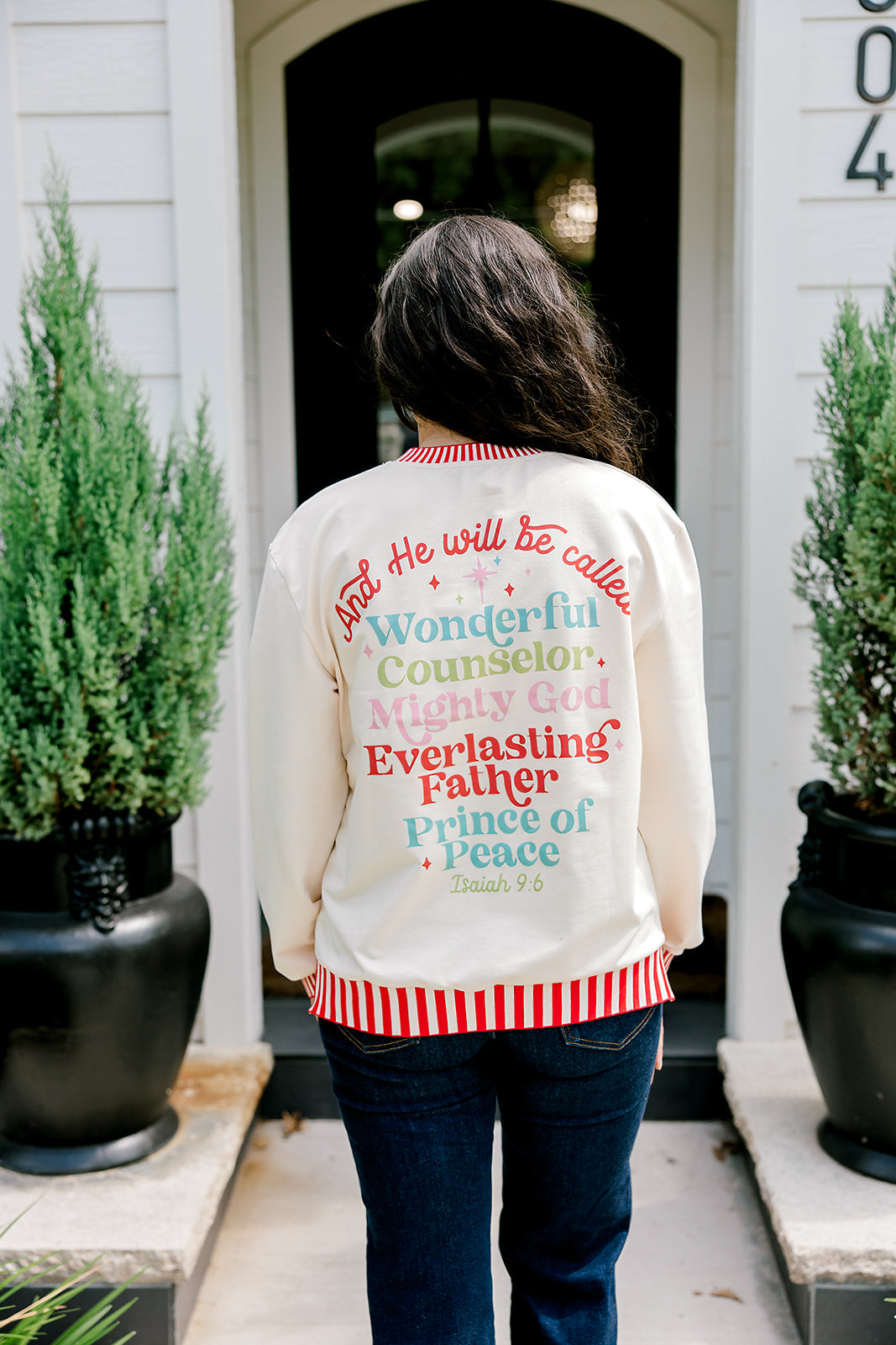 Woman with dark hair wearing a sweatshirt with red & white striping along cuff and collar. On the back of top there is a multi color bible verse. Model paired this top with dark-wash denim jeans. 