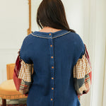 Woman with dark hair standing in a back profile position wearing the Tammy Top in a lifestyle scene. The focal point of this image is the buttons down back of top as well as the patch-work ruffled sleeves. The model paired this top with mid-wash denim jeans.