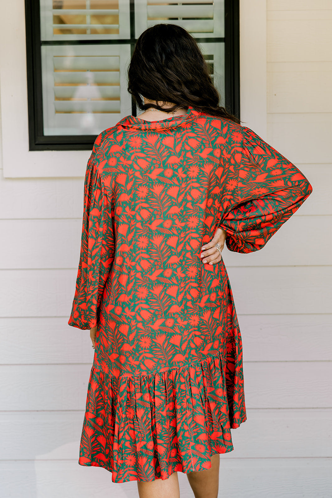 Woman with dark hair wearing a red/green otomi graphic dress that is long-sleeve & has slight ruffle at bottom of dress.