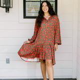 Woman with dark hair wearing a red/green otomi graphic dress that is long-sleeve & has slight ruffle at bottom of dress.
