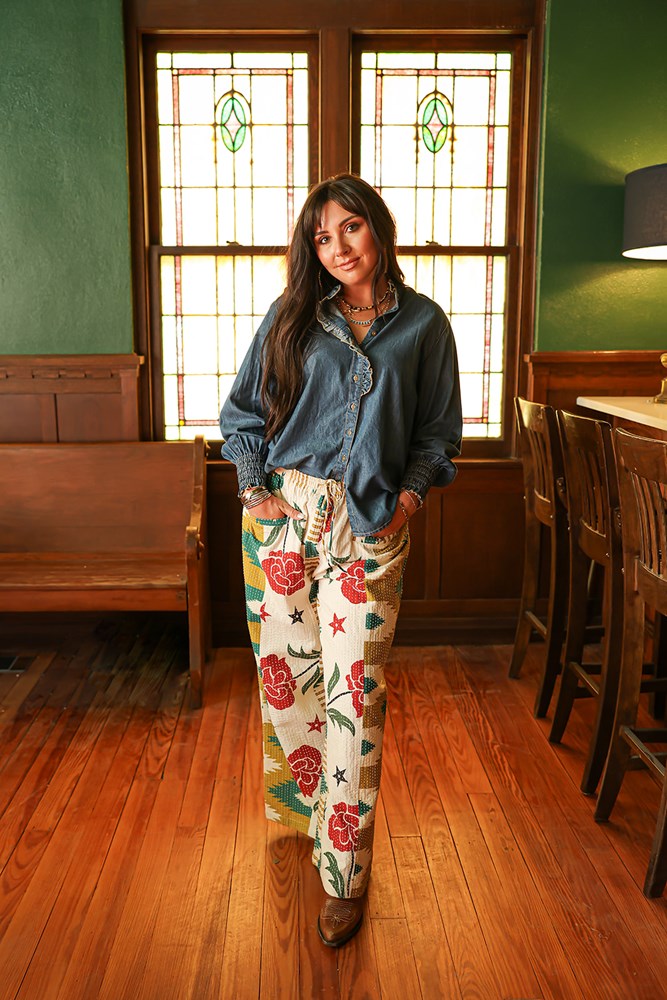 Woman with dark hair wearing a denim long-sleeve with cuffed sleeves and slight ruffling along button-line & aztec, rose, and star pants on a white background. 