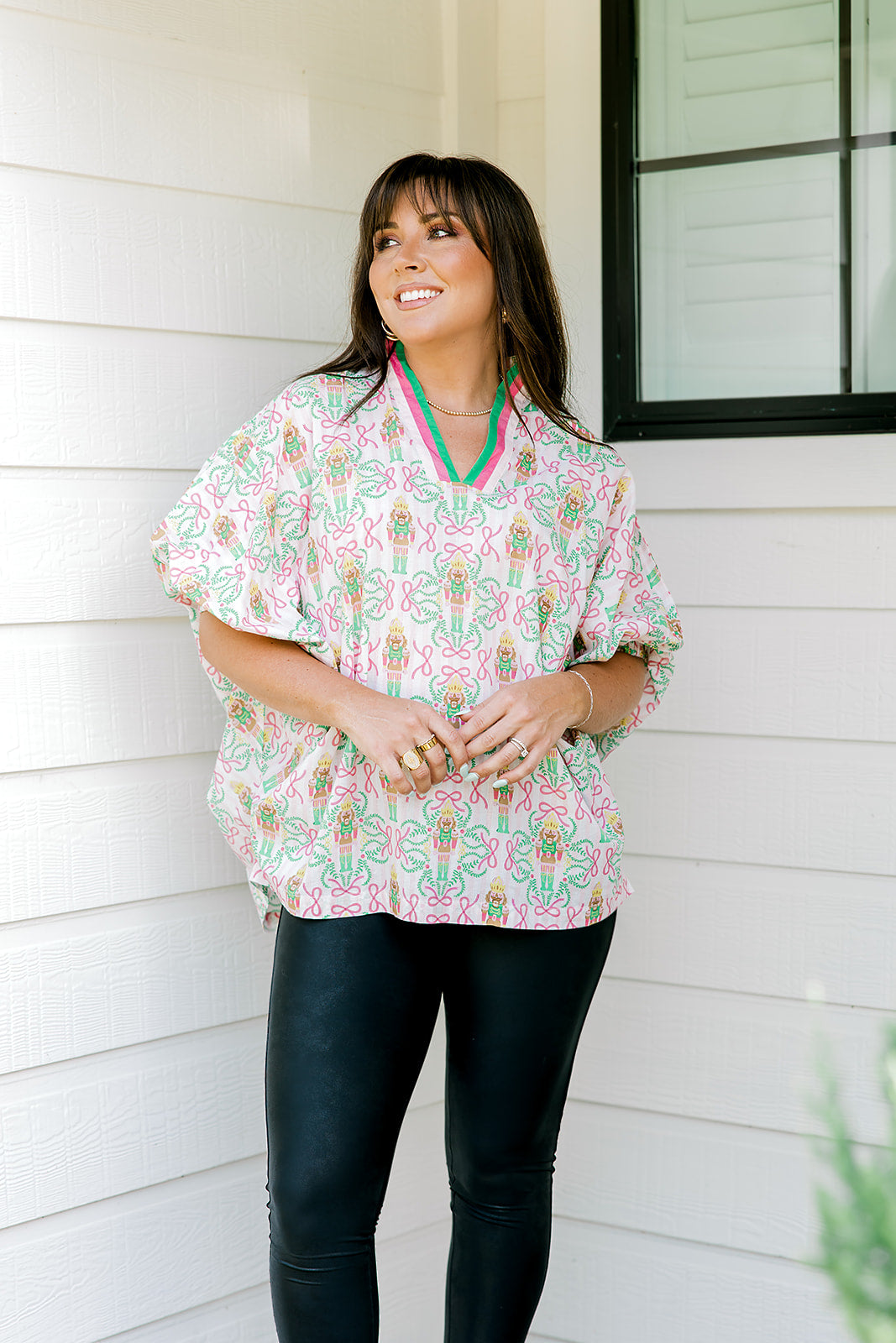 Woman with dark hair wearing a top in the Tori body style featuring pink & green nutcracker graphics. Model paired this top with faux leather leggings.