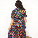 Woman with dark hair in a back profile position wearing the Vanessa Dress against a while background. The focal point of this image is the graphics on the back of the dress with small emphasis on belt and back of sleeves. 