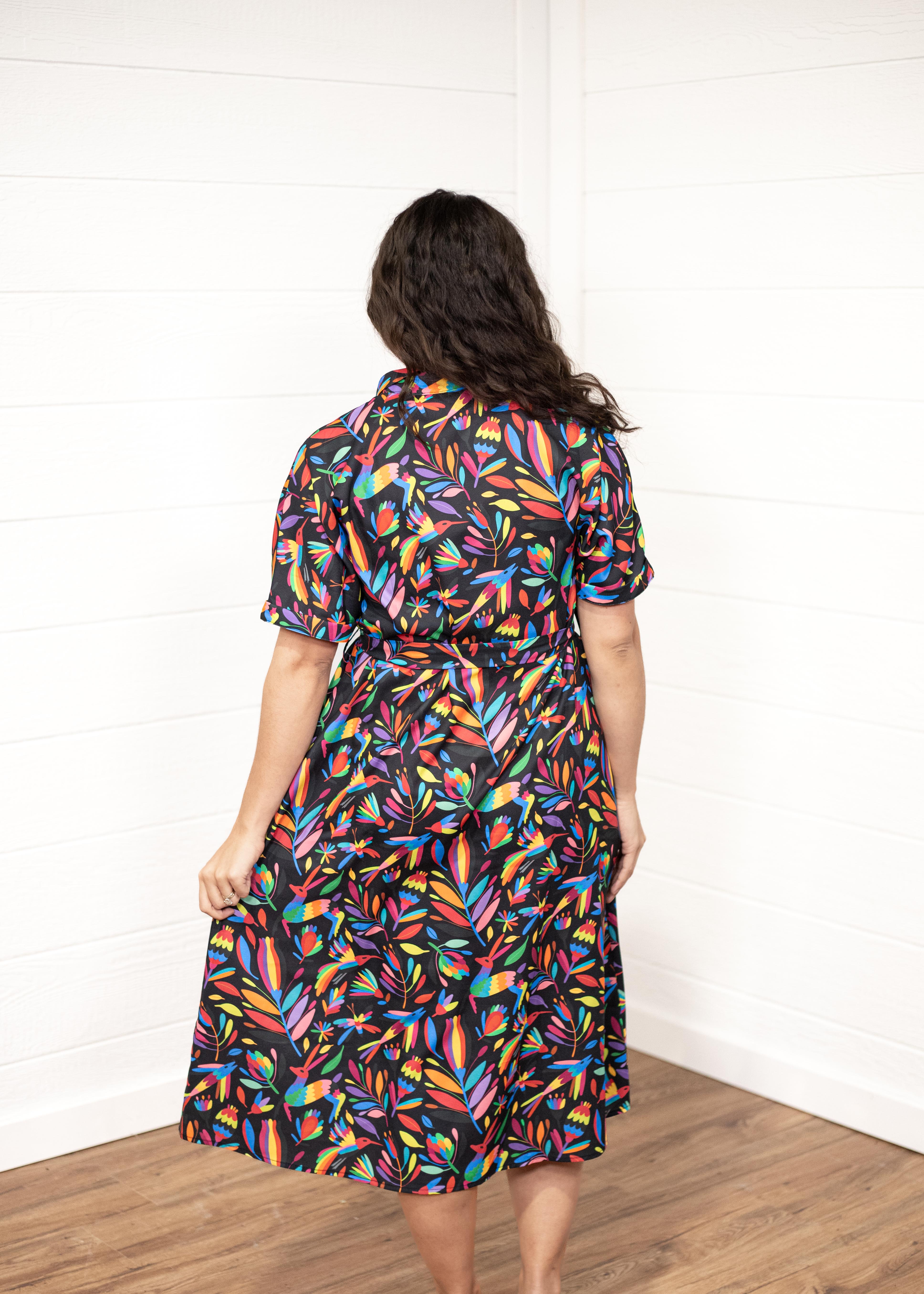 Woman with dark hair in a back profile position wearing the Vanessa Dress against a while background. The focal point of this image is the graphics on the back of the dress with small emphasis on belt and back of sleeves. 