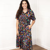 Woman with dark hair in front profile position wearing the Vanessa Dress against a white background. The focal point of this image is the front graphics on the dress with emphasis on the collar and belt. Model paired white sandals with this dress.
