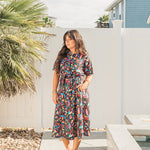 Woman with dark hair standing in front profile position wearing the Vanessa Dress in a lifestyle themed background. The focal point of this image is the entirety of the dress with bold and bright colors in the background. The model paired this dress with red sneakers.