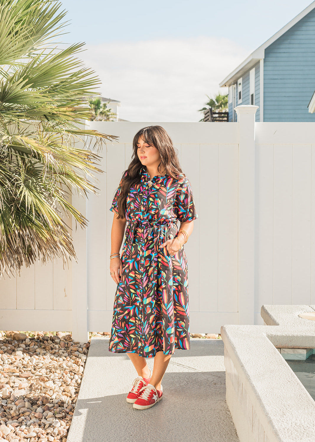 Woman with dark hair standing in front profile position wearing the Vanessa Dress in a lifestyle themed background. The focal point of this image is the entirety of the dress with bold and bright colors in the background. The model paired this dress with red sneakers.