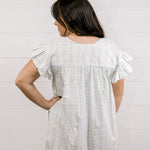 Woman wearing a white dress with ruffled sleeves against a light background facing back. 
