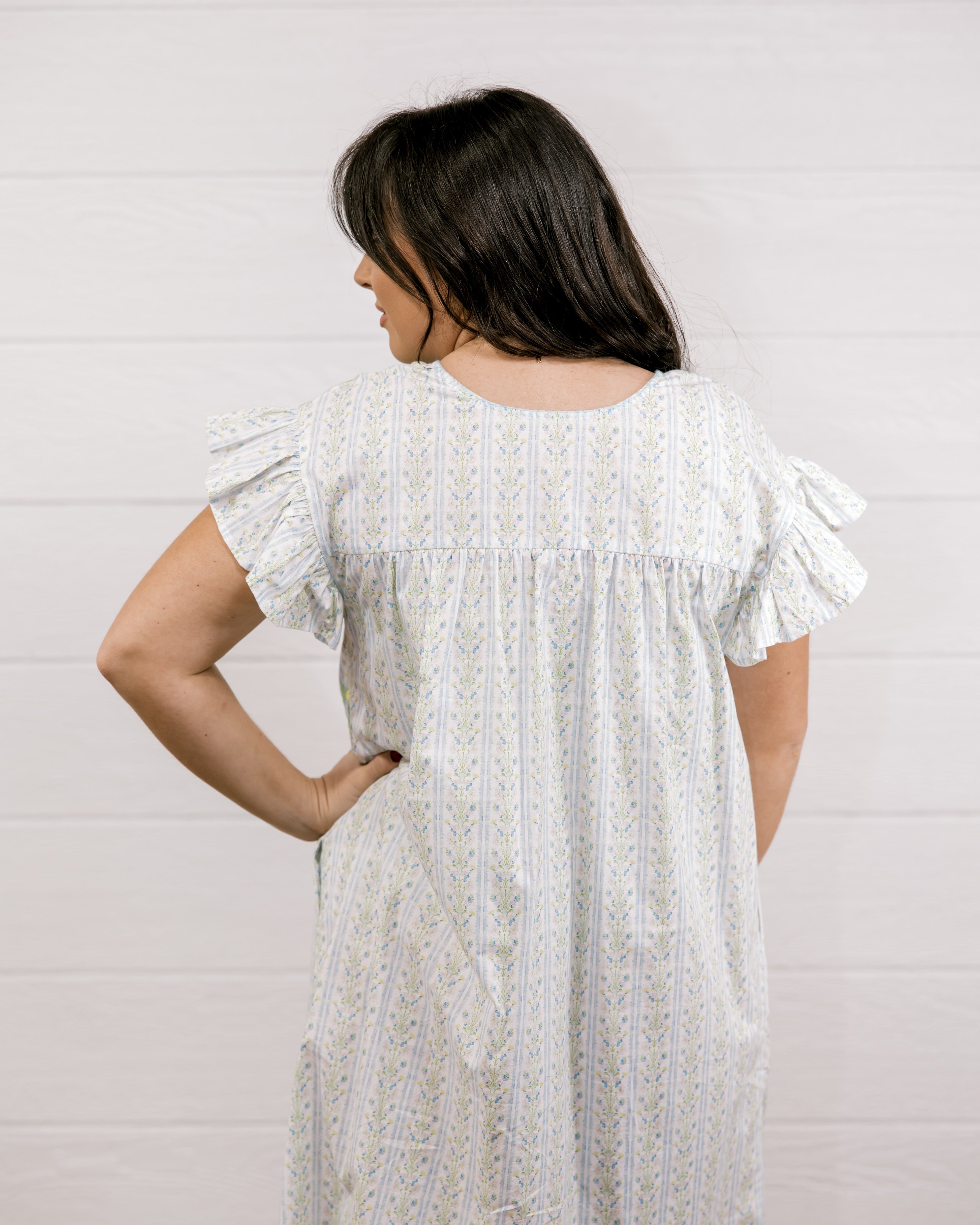 Woman wearing a white dress with ruffled sleeves against a light background facing back. 