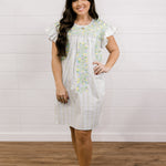 Woman wearing a white dress with floral patterns against a white wooden panel background facing front. 