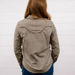 A woman viewed from behind wearing a brown and beige houndstooth patterned long-sleeve shirt with a western-style curved seam detail on the upper back, paired with blue jeans, standing against a white shiplap wall.