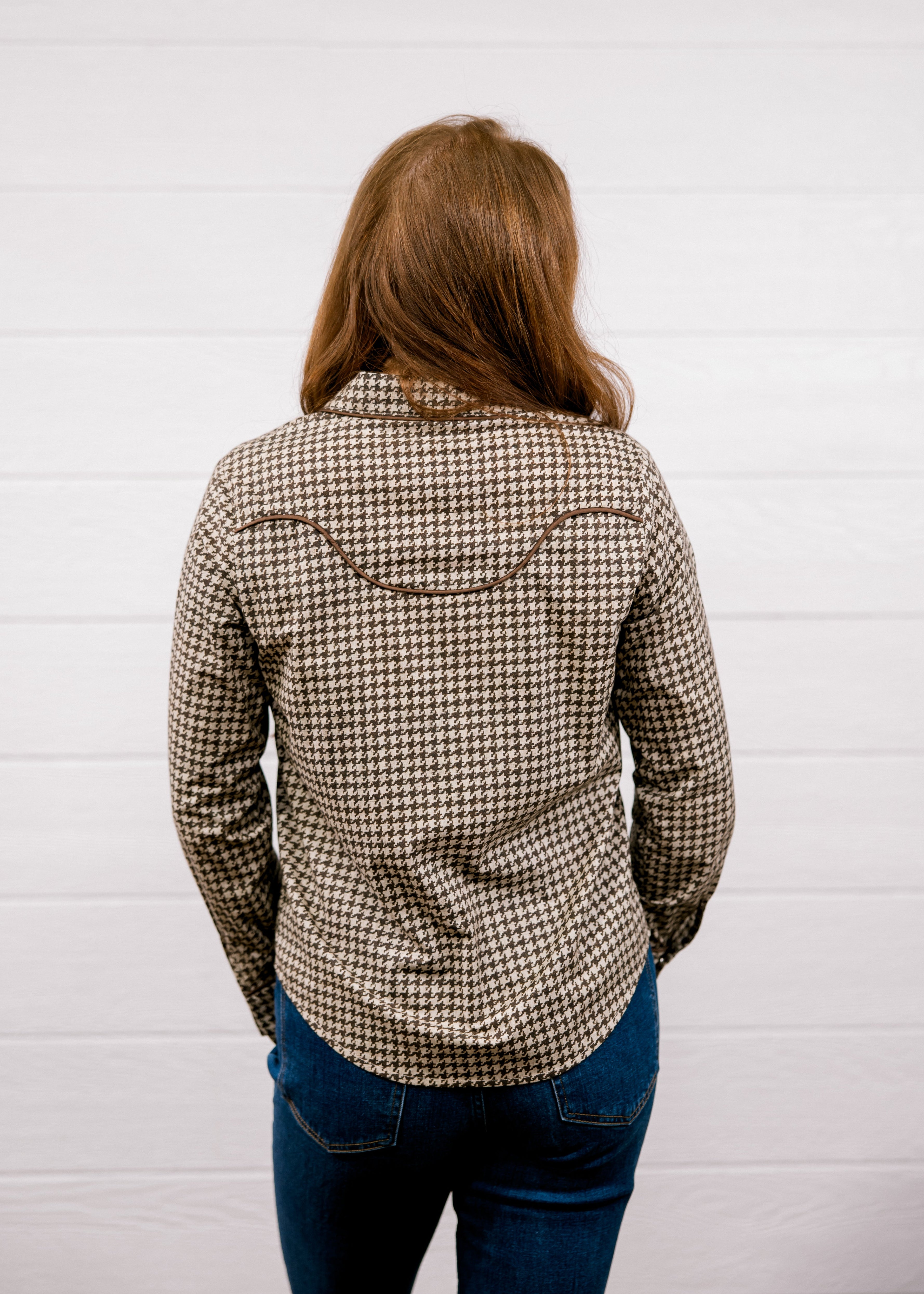 A woman viewed from behind wearing a brown and beige houndstooth patterned long-sleeve shirt with a western-style curved seam detail on the upper back, paired with blue jeans, standing against a white shiplap wall.