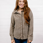 A smiling young woman with long light brown hair stands against a white wood plank wall, wearing a brown and black houndstooth button-up shirt with dark blue jeans.