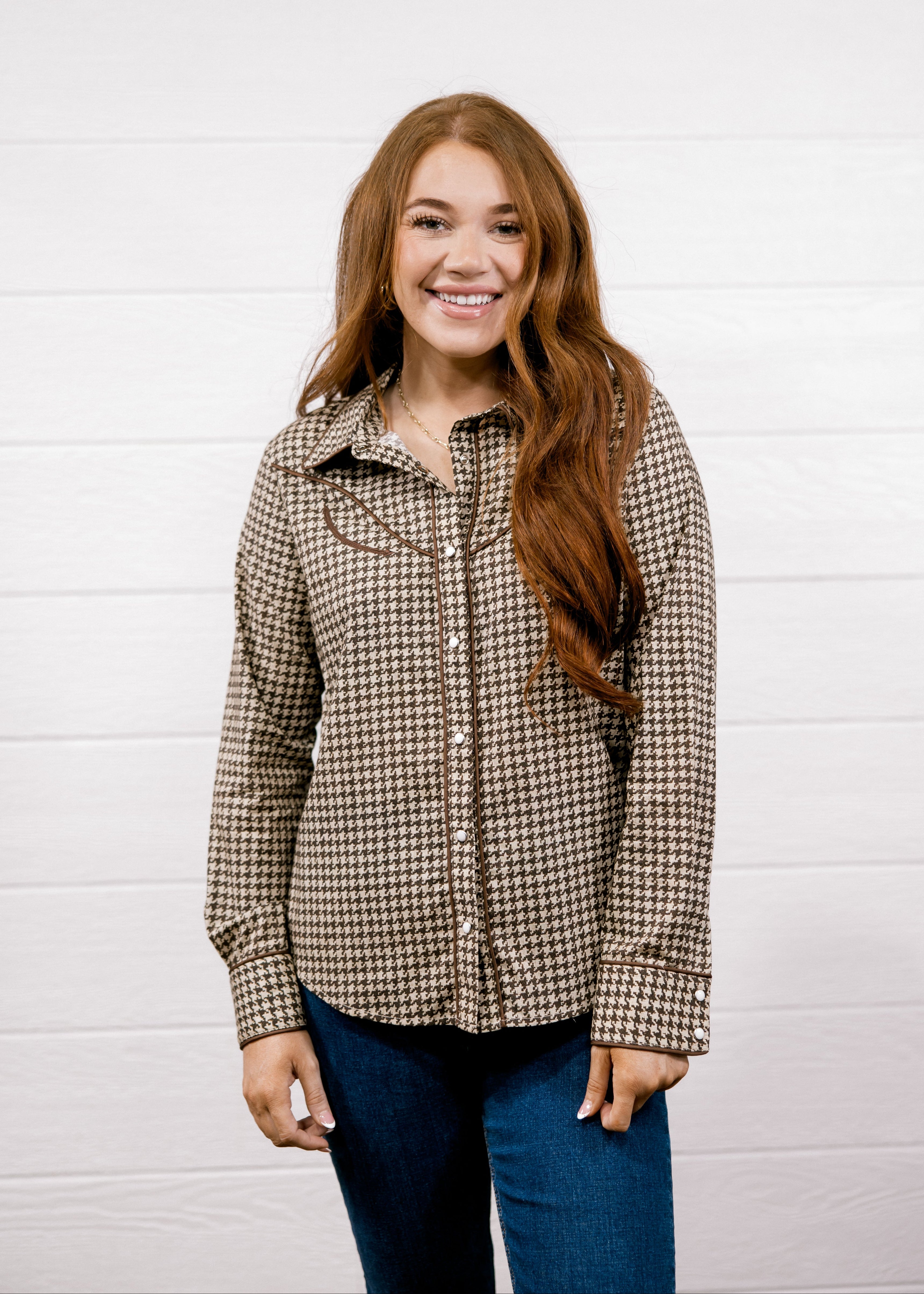 A smiling young woman with long light brown hair stands against a white wood plank wall, wearing a brown and black houndstooth button-up shirt with dark blue jeans.