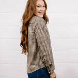 A woman with long wavy red hair is shown from the back and side, wearing a brown and beige houndstooth patterned button-up shirt and dark blue jeans, standing against a white wood-paneled wall.