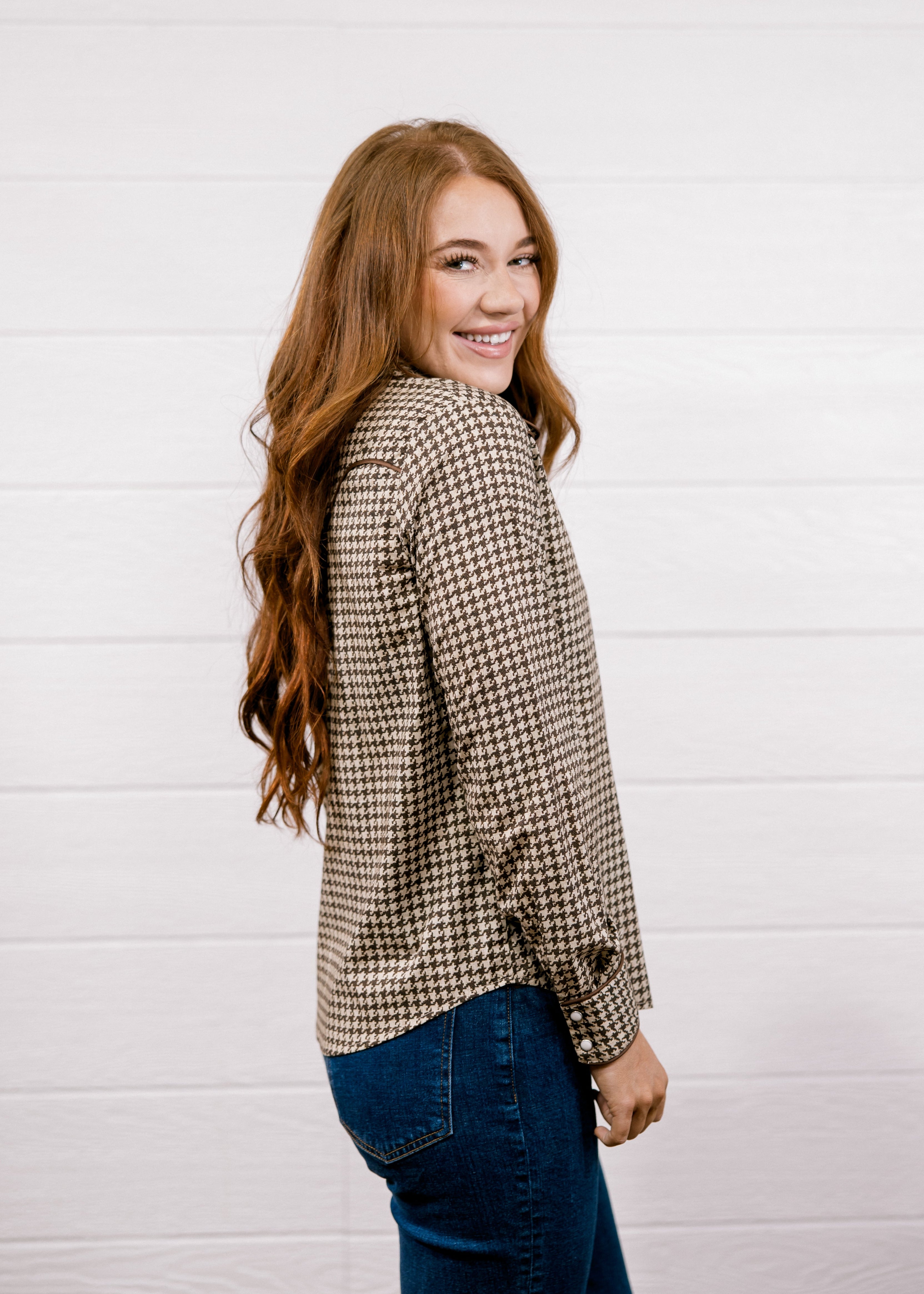 A woman with long wavy red hair is shown from the back and side, wearing a brown and beige houndstooth patterned button-up shirt and dark blue jeans, standing against a white wood-paneled wall.