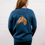 A woman viewed from behind wearing a navy blue crewneck sweater with a detailed brown horse head graphic on the back, paired with dark blue jeans, set against a white wood-paneled wall.