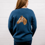 A woman viewed from behind wearing a navy blue crewneck sweater with a detailed brown horse head graphic on the back, paired with dark blue jeans, set against a white wood-paneled wall.
