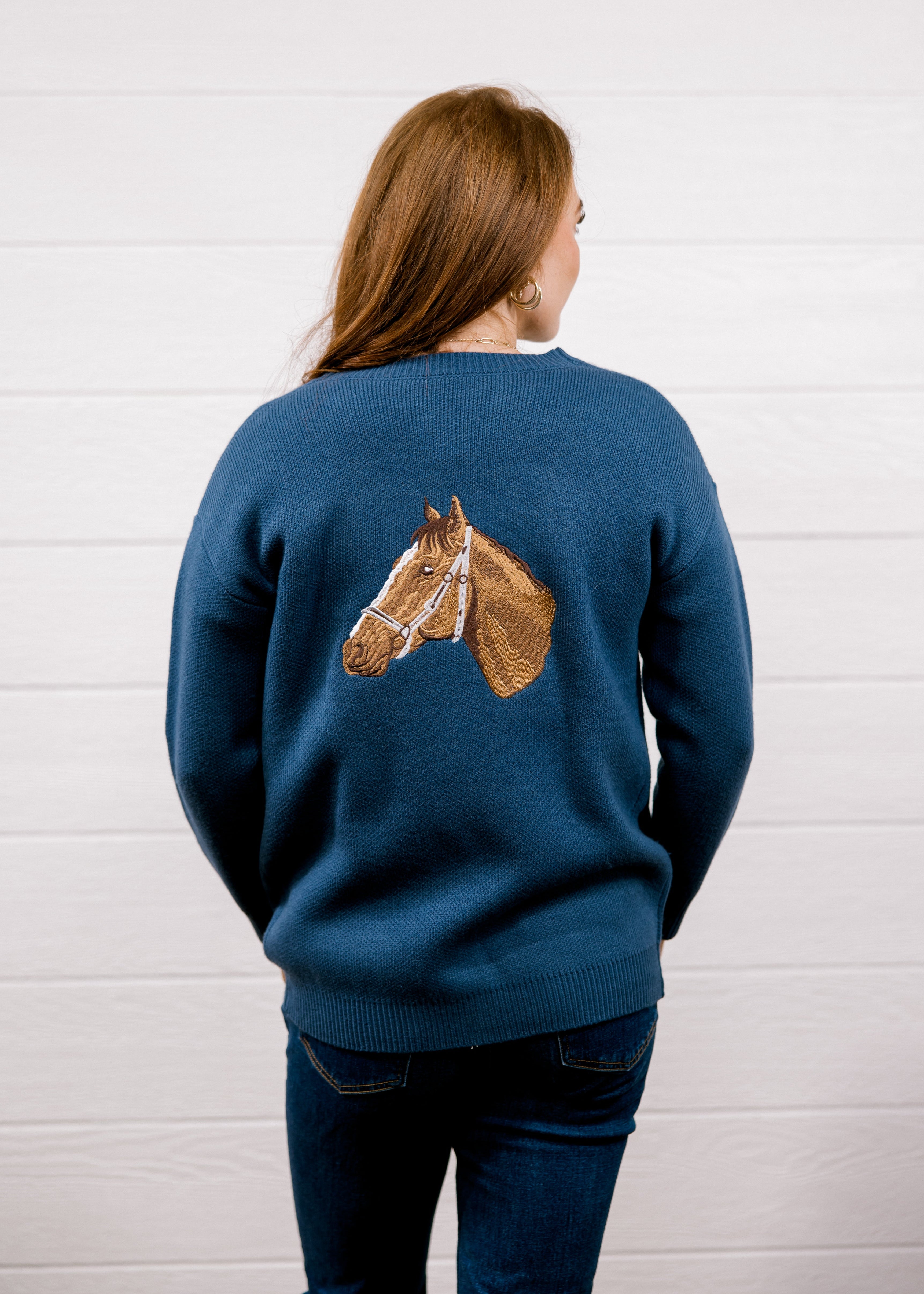 A woman viewed from behind wearing a navy blue crewneck sweater with a detailed brown horse head graphic on the back, paired with dark blue jeans, set against a white wood-paneled wall.