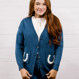 A woman with long red hair stands against a white wood-paneled wall wearing a blue cardigan with white horseshoe details on the pockets, paired with matching blue jeans and a white top.