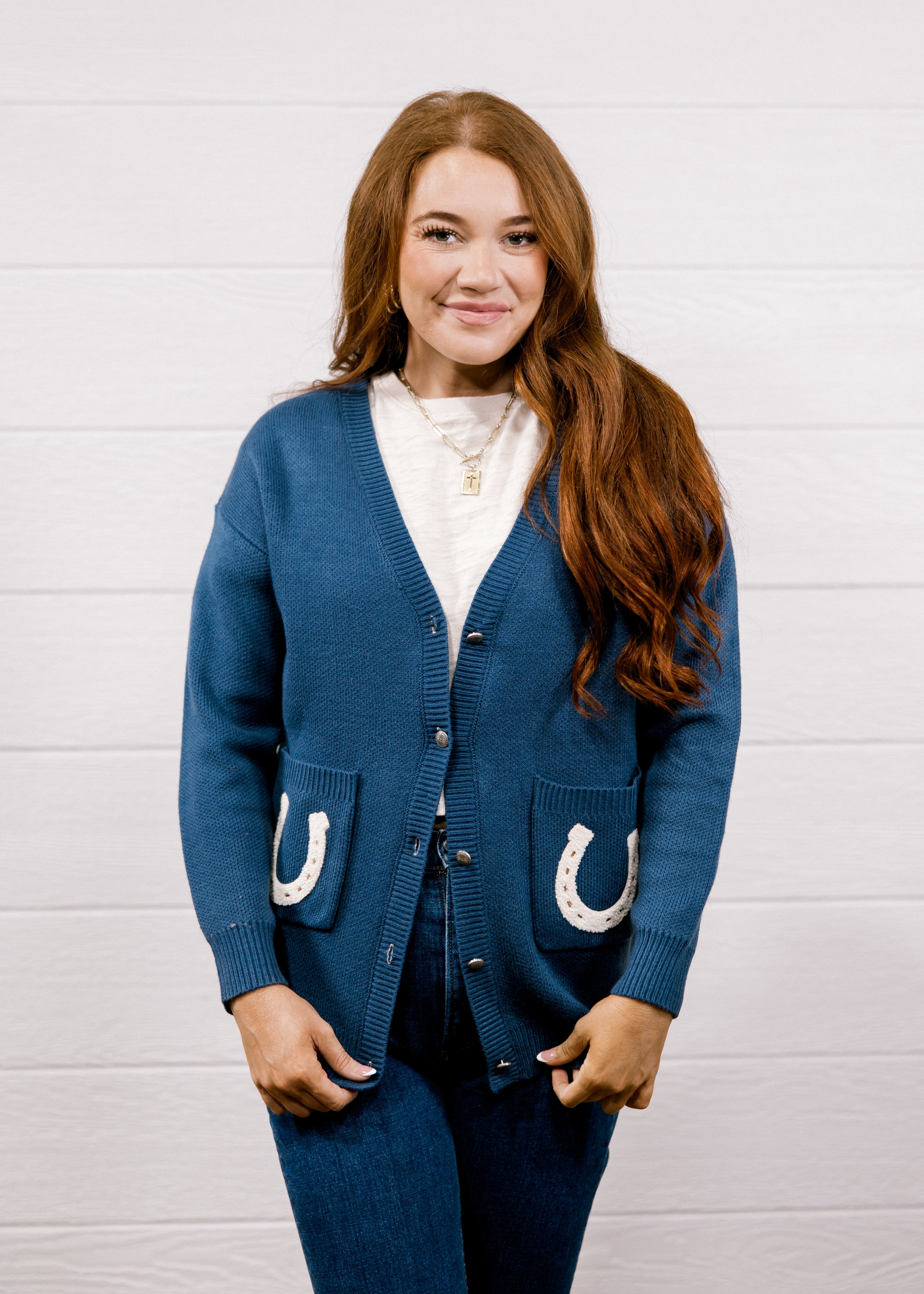 A woman with long red hair stands against a white wood-paneled wall wearing a blue cardigan with white horseshoe details on the pockets, paired with matching blue jeans and a white top.