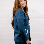 A woman with long light brown hair is shown from the side and back, wearing a blue knit cardigan with a white crescent moon detail on the sleeve, paired with dark blue jeans, against a white wood-paneled wall.
