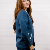 A woman with long light brown hair is shown from the side and back, wearing a blue knit cardigan with a white crescent moon detail on the sleeve, paired with dark blue jeans, against a white wood-paneled wall.