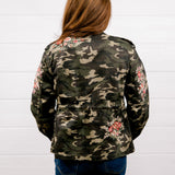 A woman viewed from behind wearing a camouflage military-style jacket with pink floral embroidery on the sleeves and back, paired with blue jeans, standing against a white wood-paneled wall.